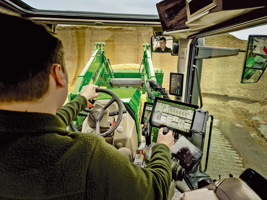 Pogled na unutrašnjost kabine traktora John Deere, koji prikazuje komande rukovaoca i rad prednjeg utovarivača.