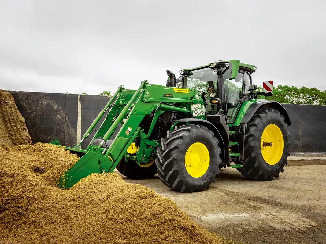 Traktor John Deere koji se prednjom kašikom utovarivača zabija u gomilu silaže u poljoprivrednom okruženju.