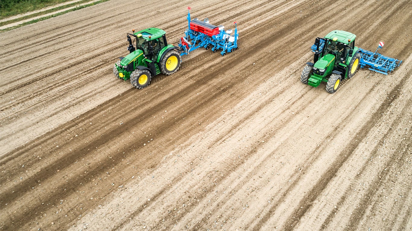 Dva John Deere 6M traktora u golom polju. Jedan vuče Monsomen sadilicu. Drugi vuče Lemken kombinaciju za pravljenje leja