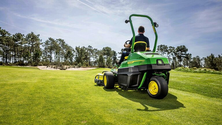 Čovek vozi John Deere 2775 E-Cut™ Electric kosačicu za travnate terene dobro održavanim terenom za golf, sa drvećem i delimično oblačnim nebom u pozadini.