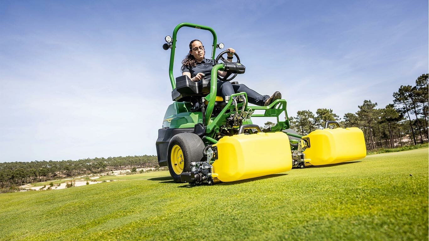 Čovek vozi zelenu i žutu John Deere 2775 E-Cut™ Electric kosačicu sa prednjim valjcima sa žutim kućištem na terenu za golf, sa drvećem i vedrim plavim nebom u pozadini.