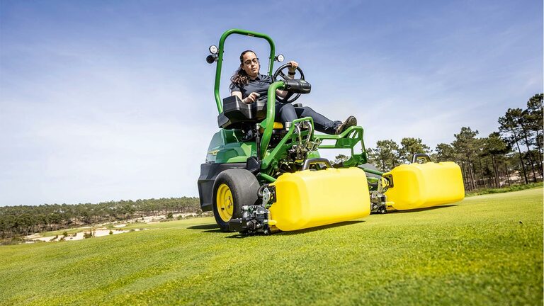 Čovek vozi zelenu i žutu John Deere 2775 E-Cut™ Electric kosačicu sa prednjim valjcima sa žutim kućištem na terenu za golf, sa drvećem i vedrim plavim nebom u pozadini.