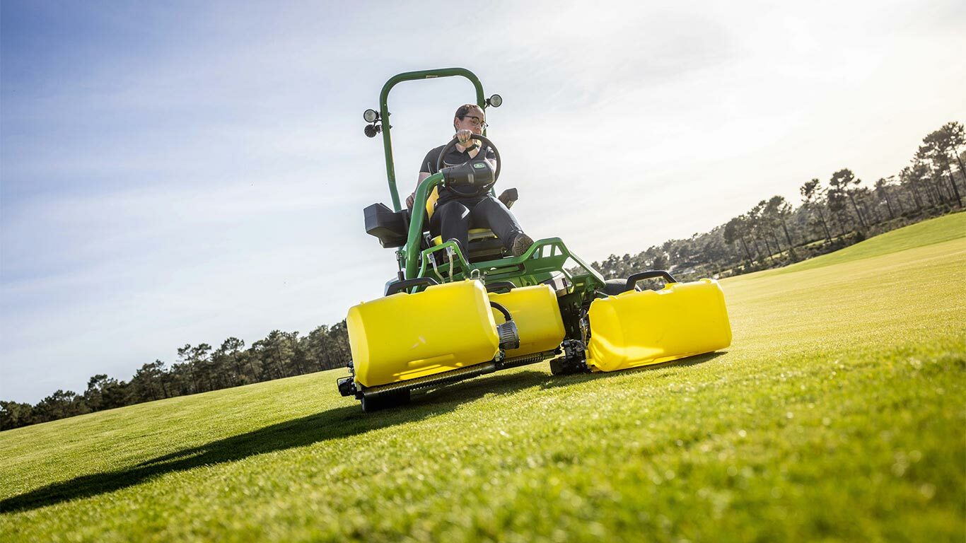 Čovek vozi zelenu i žutu John Deere 2775 E-Cut™ Electric kosačicu sa tri velika prednja valjka sa žutim kućištem na terenu za golf, sa drvećem i delimično oblačnim nebom u pozadini.
