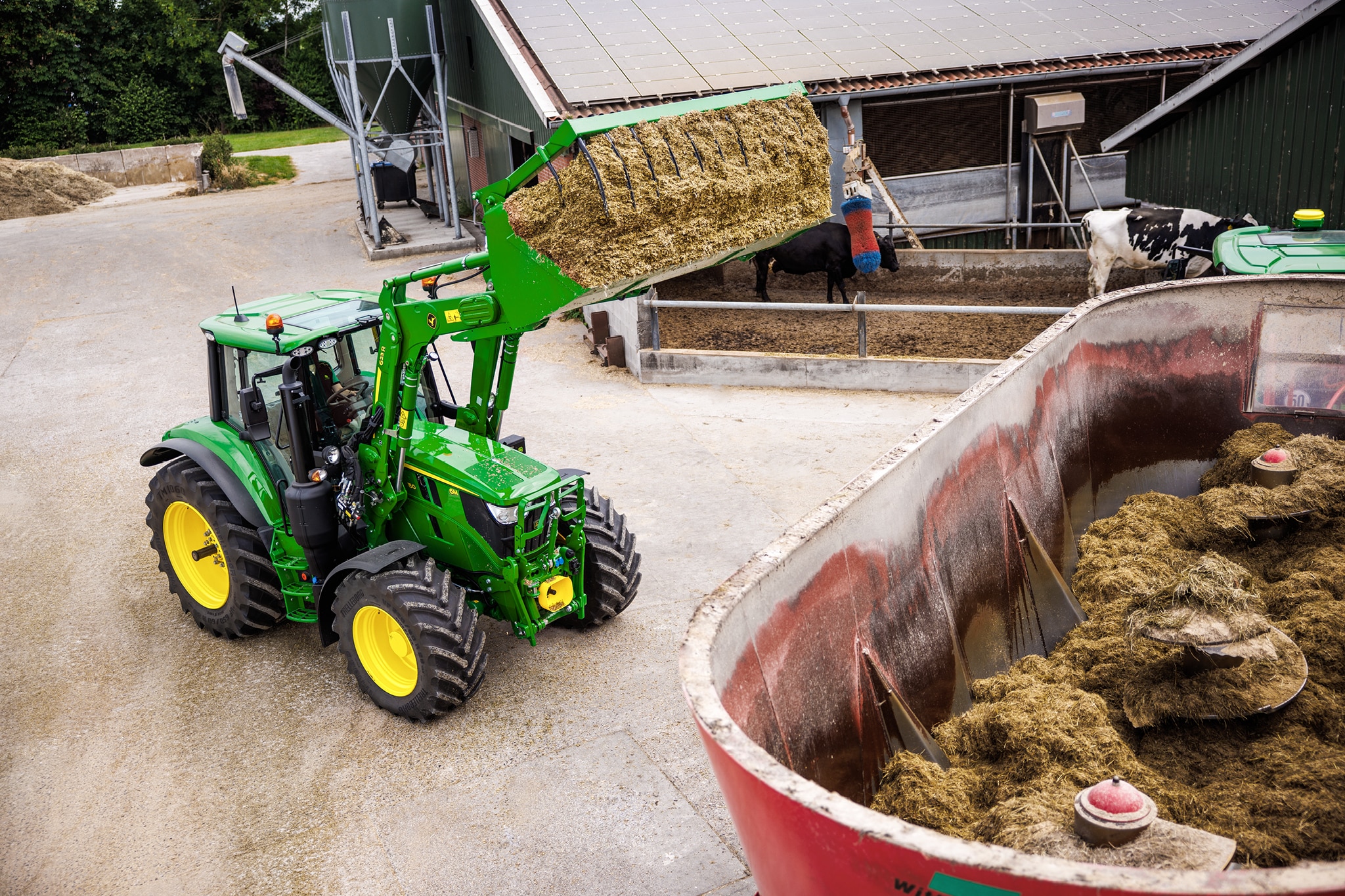 Traktor serije John Deere 6M koristi prednji utovarivač za ubacivanje silaže u mikser prikolicu na farmi.