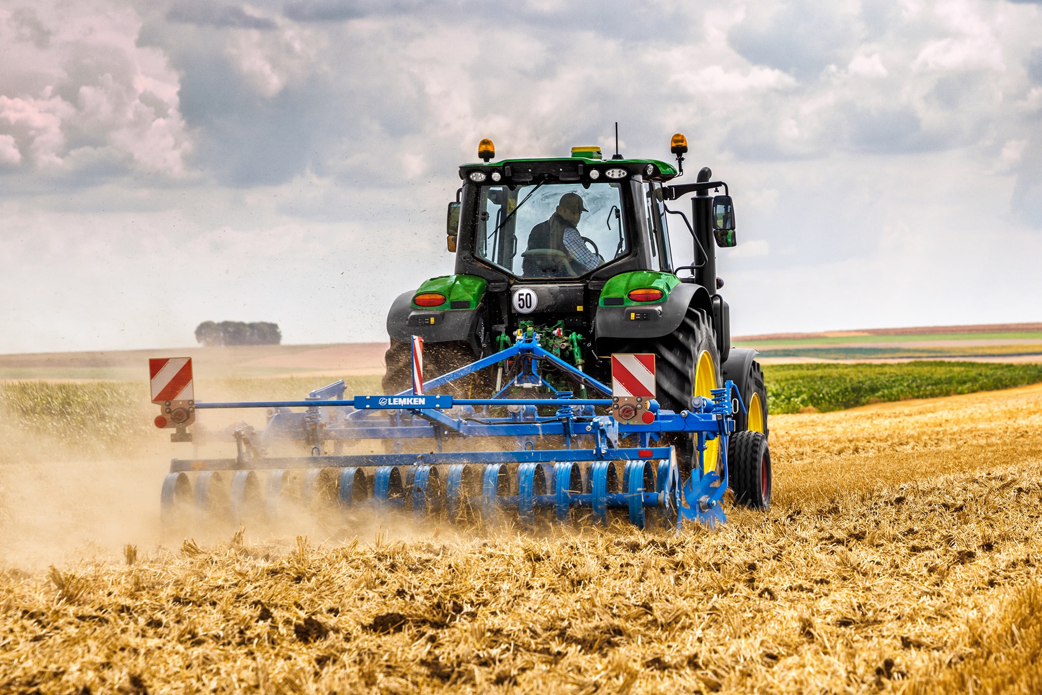 Traktor serije John Deere 6M obrađuje požnjeveno polje tanjiračom, prikaz od nazad u prašnjavim uslovima na terenu.