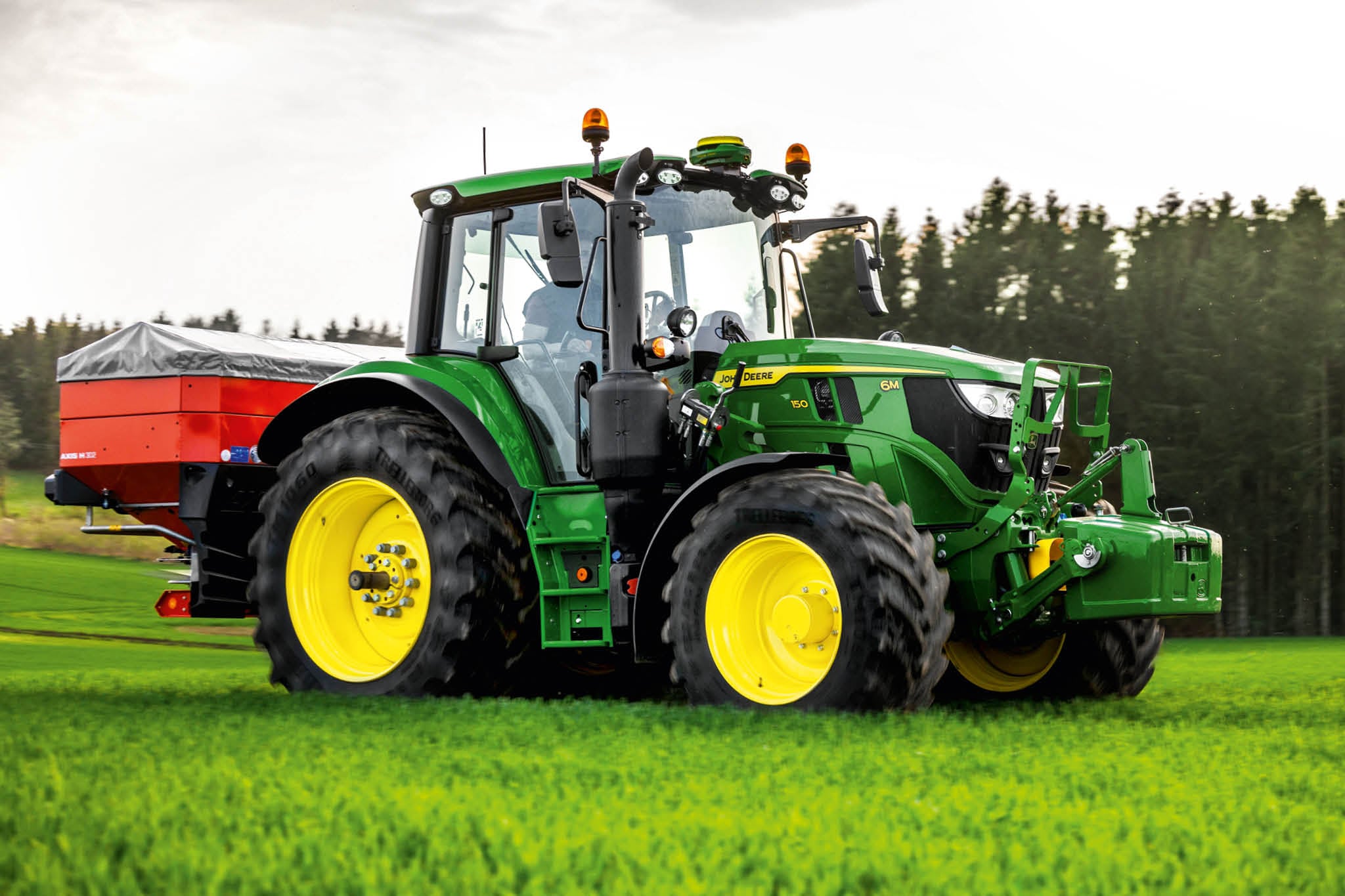 John Deere traktor serije 6M sa zadnjim rasipačem koji radi na travi blizu ruba šume.
