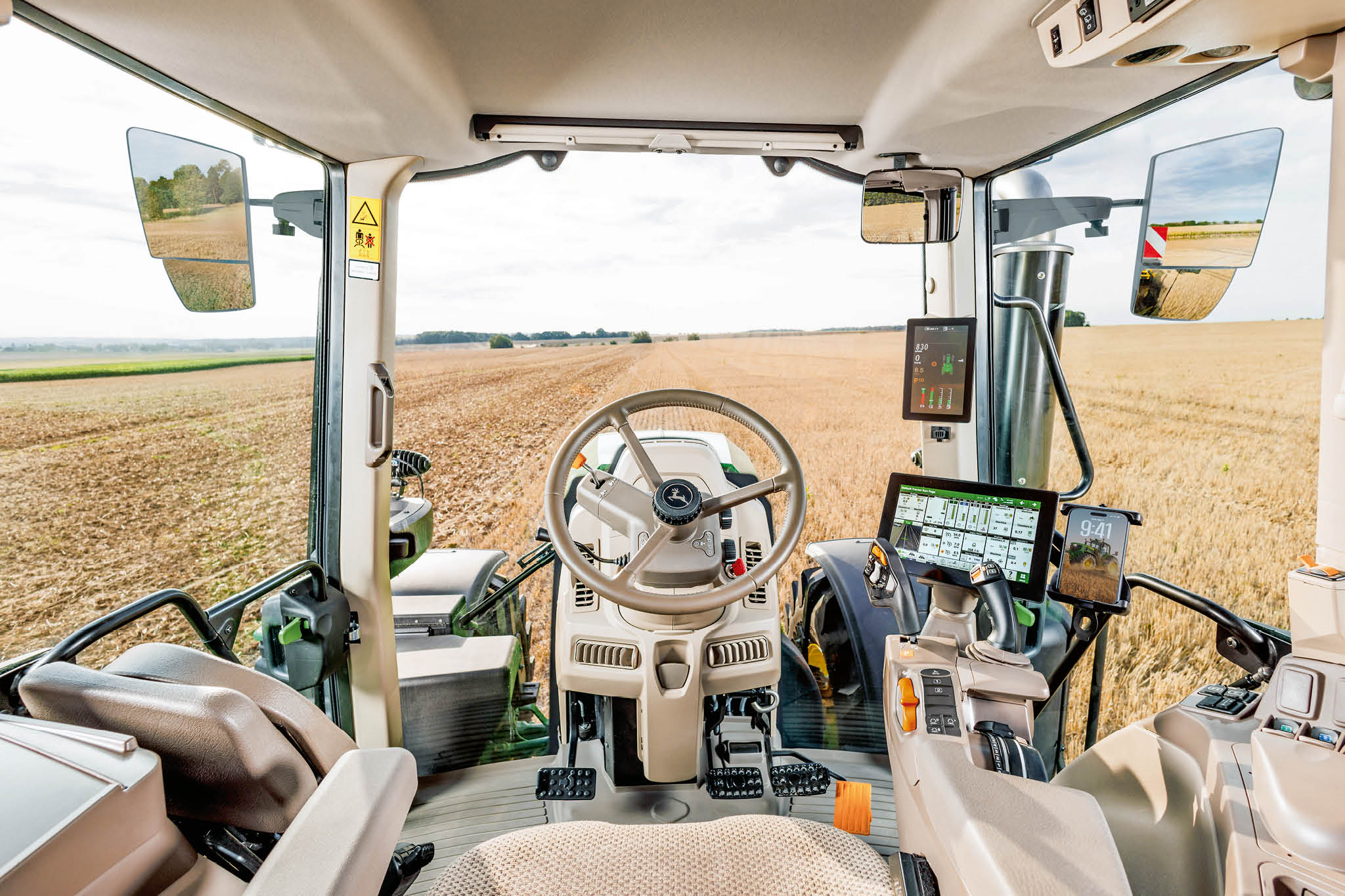 Unutrašnjost kabine traktora John Deere sa volanom, digitalnim kontrolnim ekranima i pogledom kroz vetrobransko staklo tokom rada na farmi.