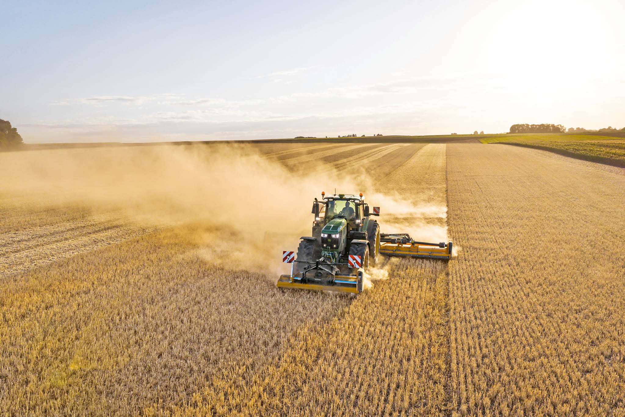 Traktor serije John Deere 6M vuče cisternu za mulj duž seoskog puta oivičenog travom i poljima.