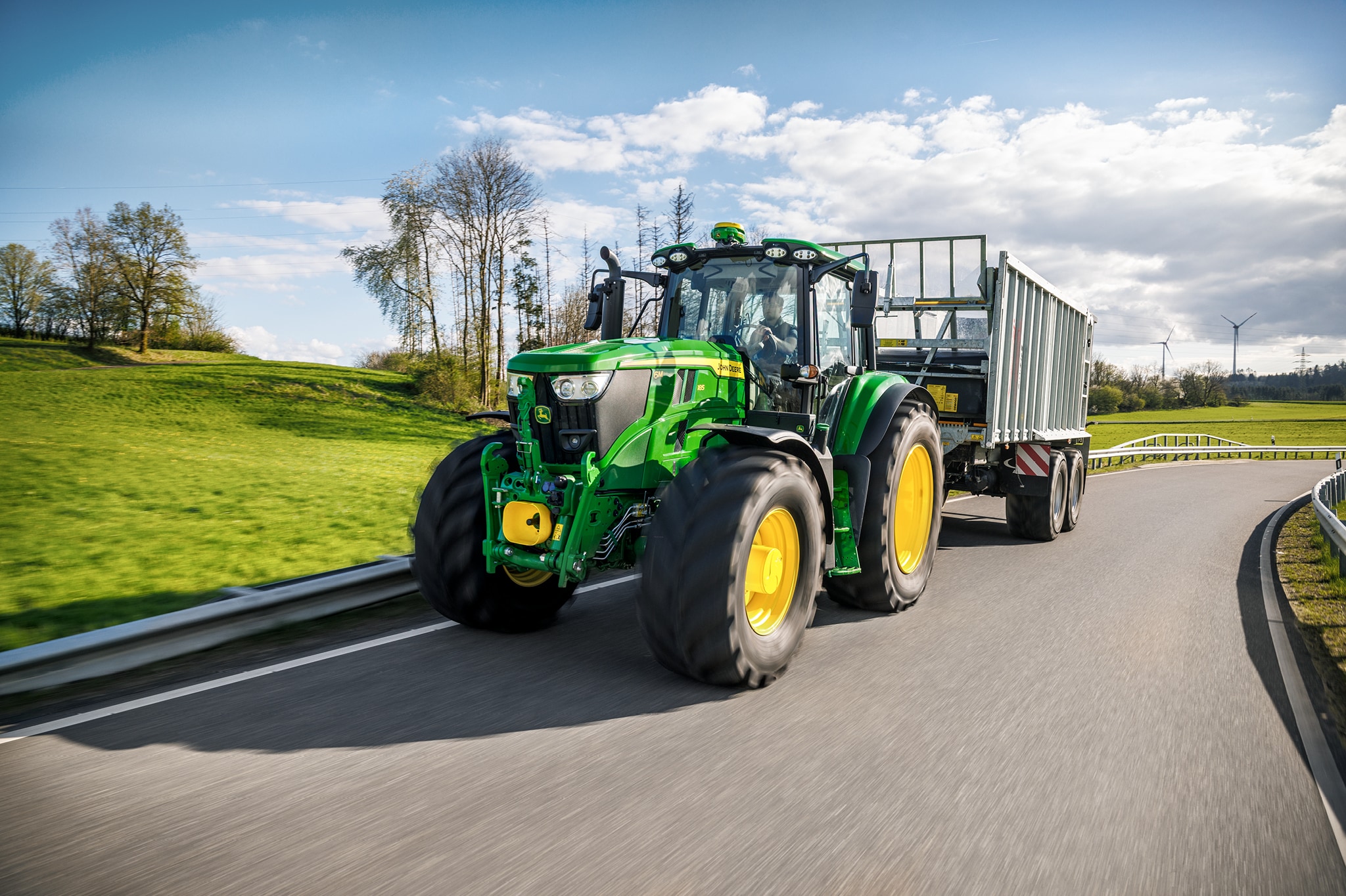 Traktor serije John Deere 6M koji prevozi prikolicu s visokim stranicama na asfaltiranom seoskom putu.