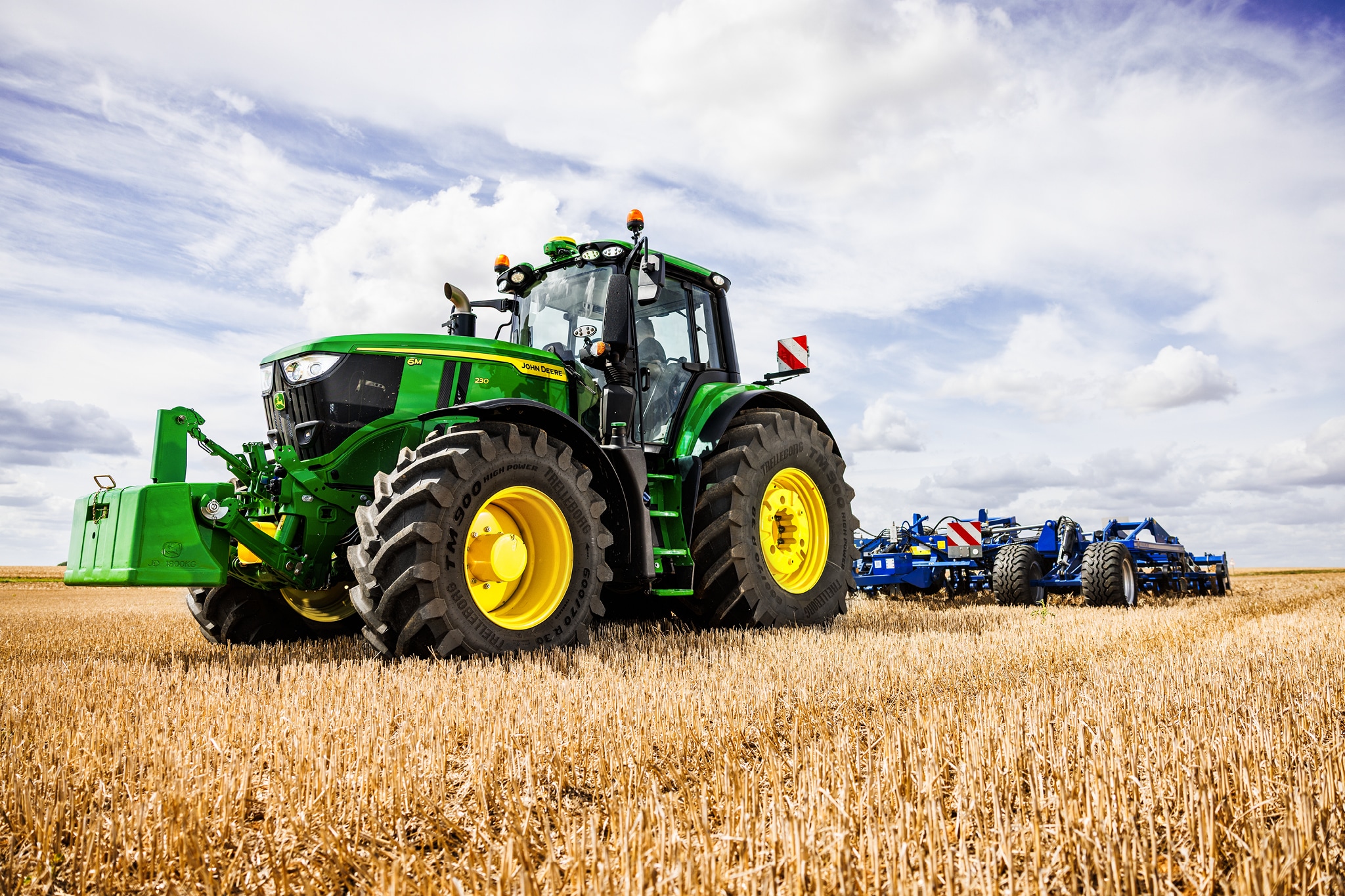 Traktor serije John Deere 6M vuče teški priključni uređaj za oranje preko strnjišta pod oblačnim nebom.