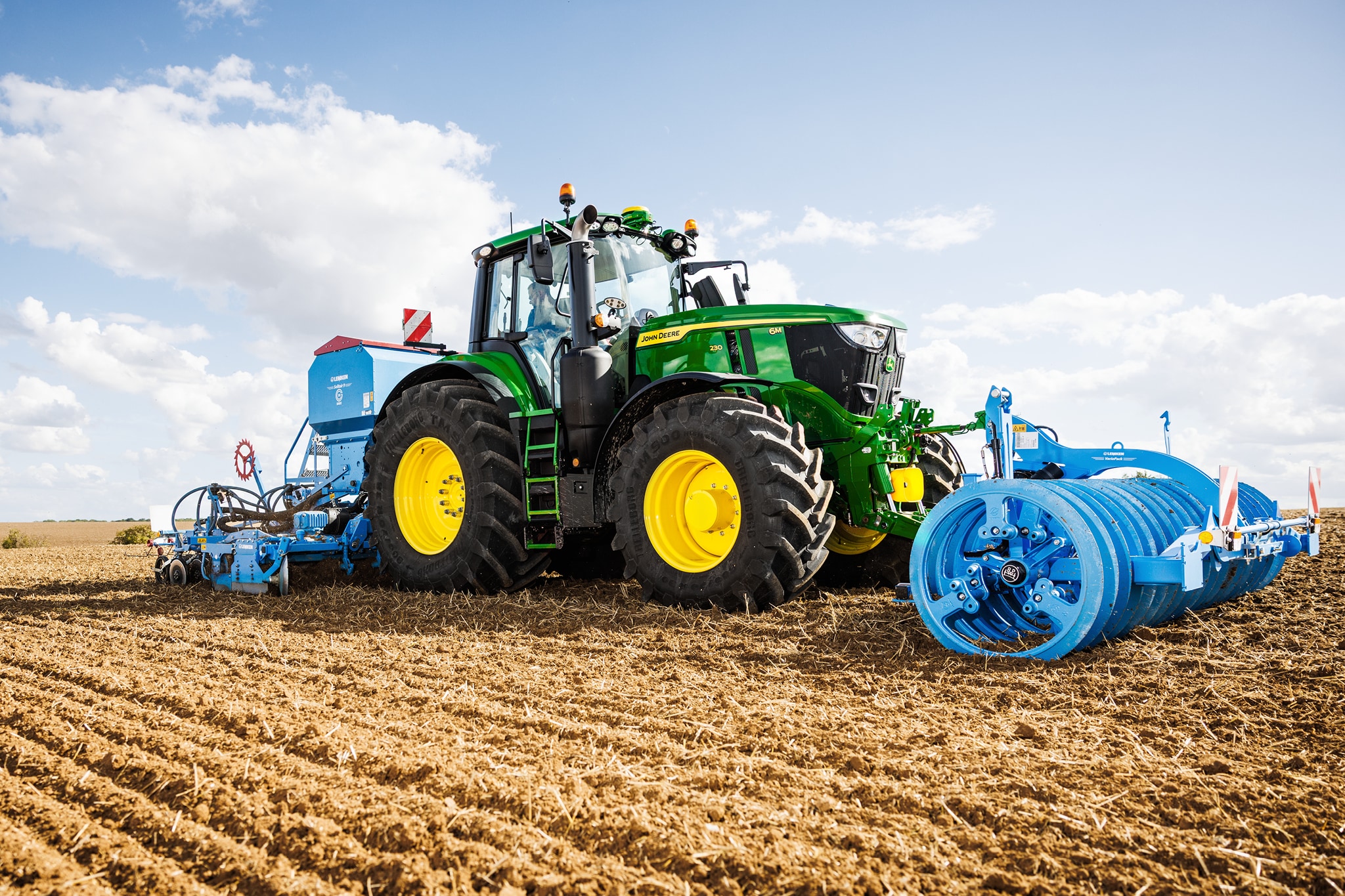 Traktor serije John Deere 6M obrađuje polje velikim plavim priključnim valjkom.