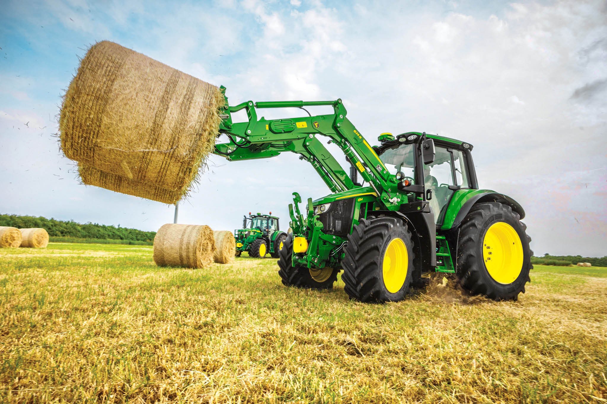Traktor serije John Deere 6M podiže veliku okruglu balu sena prednjim utovarivačem na otvorenom polju.