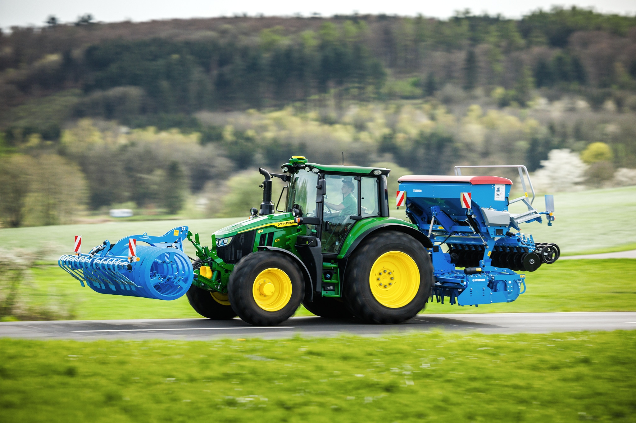 Traktor serije John Deere 6M koji prevozi priključni uređaj koji se montira napred i zadnju sejalicu duž seoskog puta.