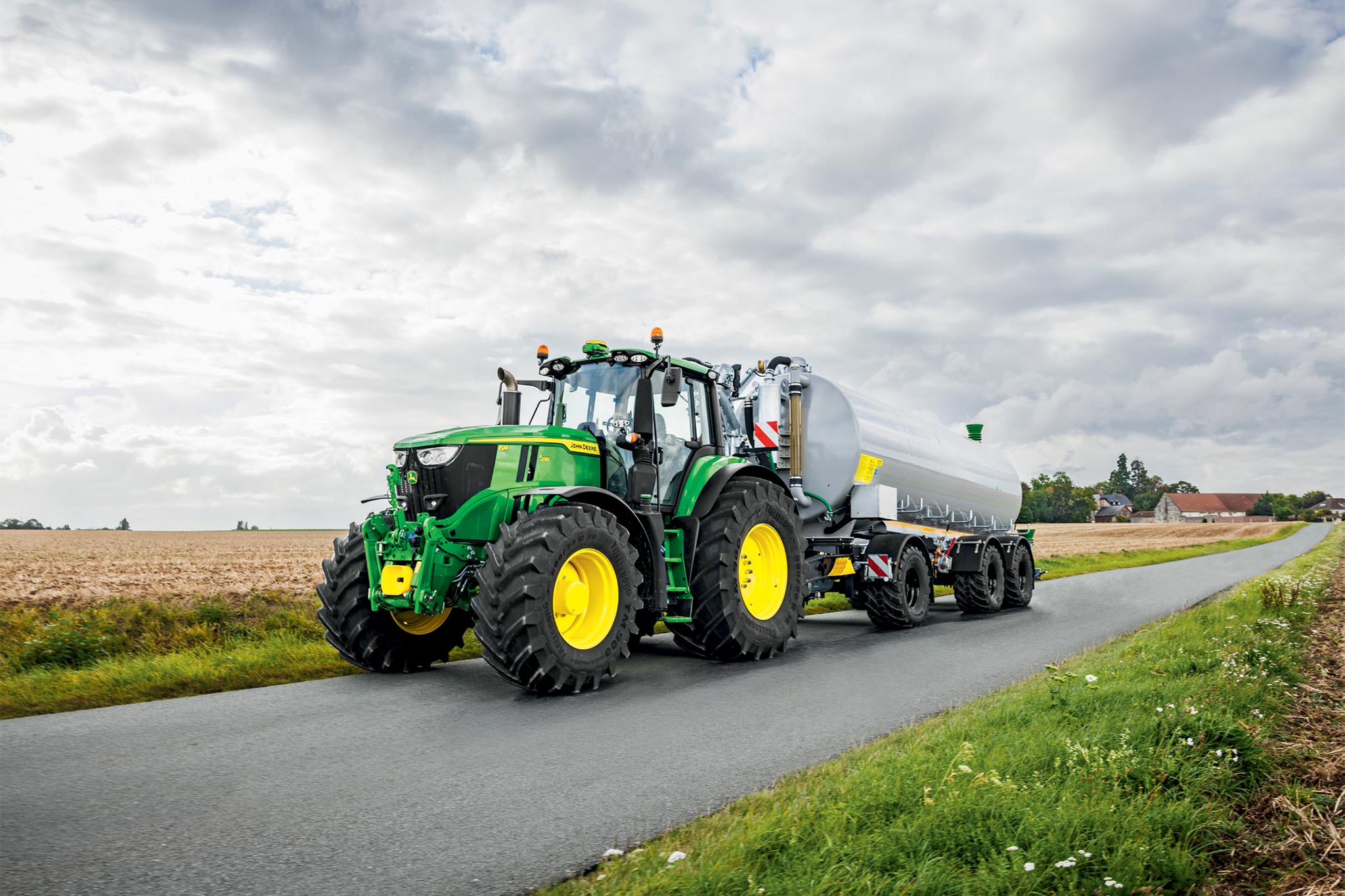 Traktor serije John Deere 6M radi na žetvenom polju sa širokim kultivatorima, gledano odozgo uz oblak prašine.