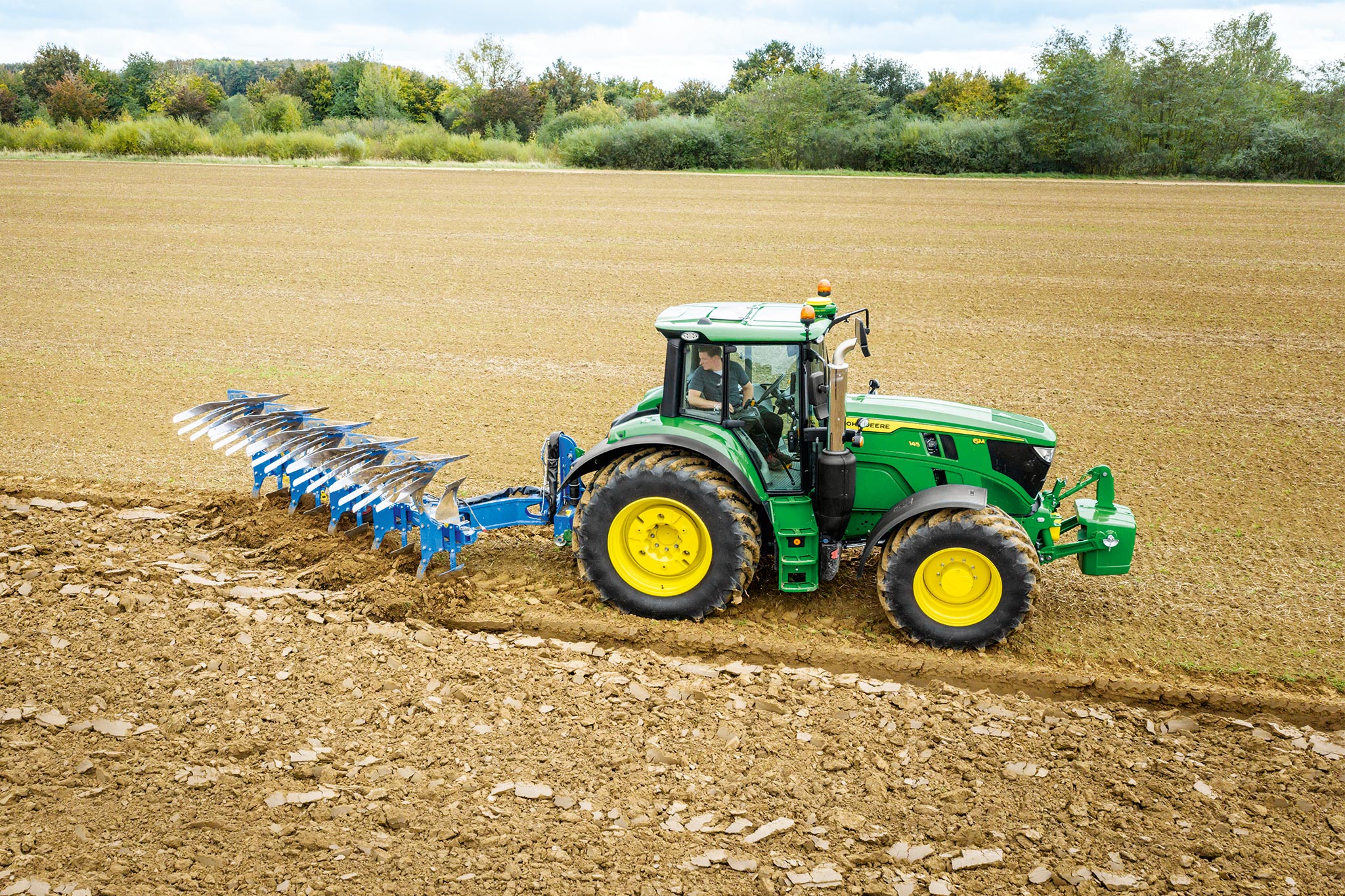 Traktor serije John Deere 6M radi na uzoranom polju sa priključnim uređajem za oranje montiranim pozadi, pod oblačnim nebom.