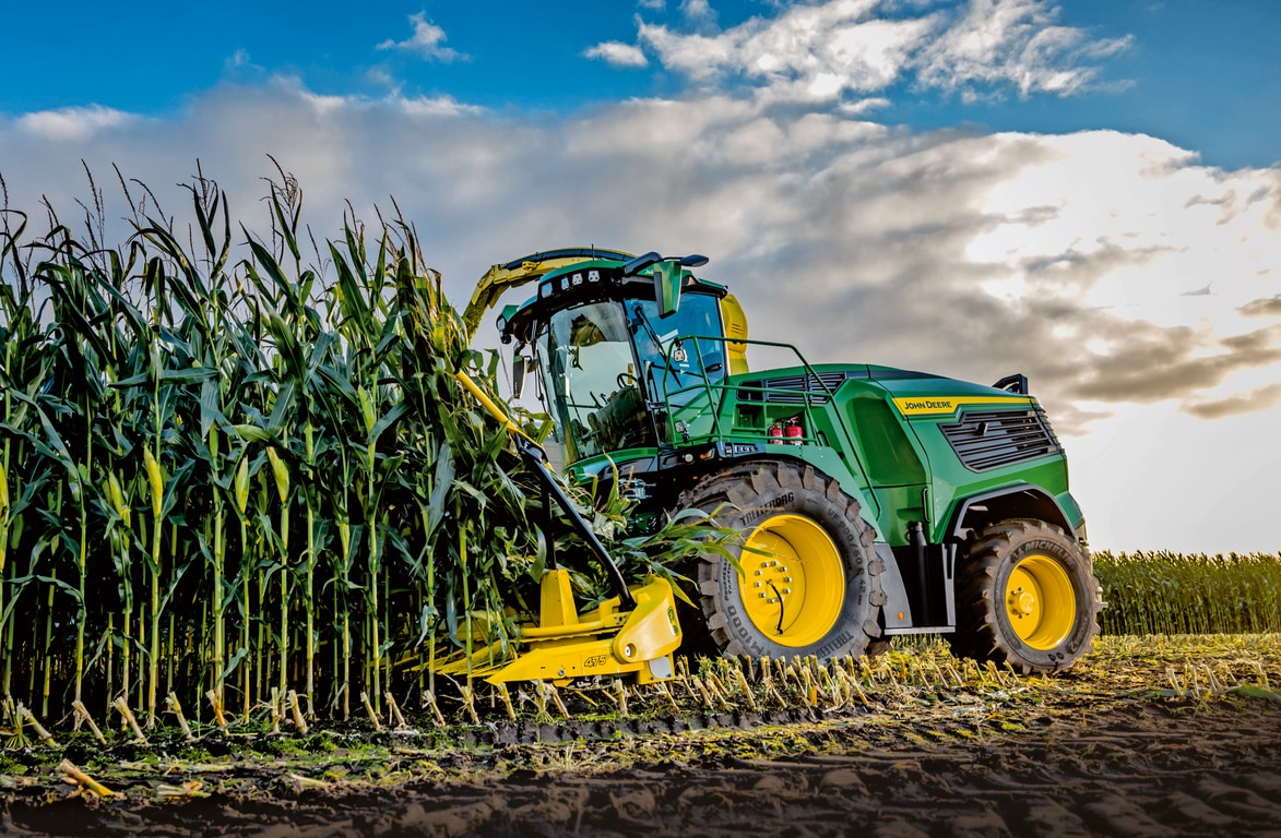 John Deere silažni kombajn žanje kukuruz na polju pod plavim nebom sa oblacima.
