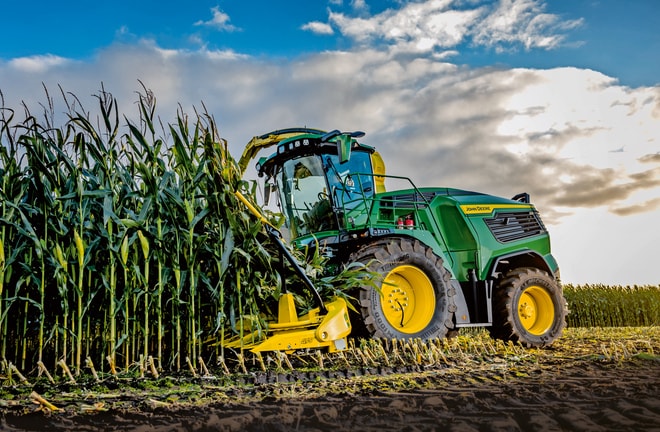 John Deere silažni kombajn žanje kukuruz na polju pod plavim nebom sa oblacima.