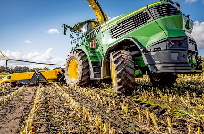 John Deere silažni kombajn radi na požnjevenom polju kukuruza, prikazuje velike pneumatike mašine i mehanizam za sečenje pod vedro plavim nebom.