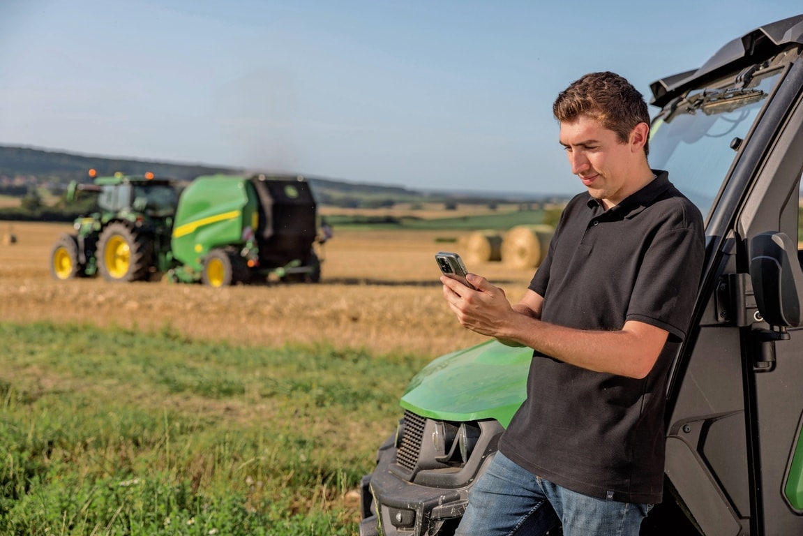 Osoba proverava podatke dokumentacije o balama na mobilnom telefonu, u pozadini je balirka John Deere VR